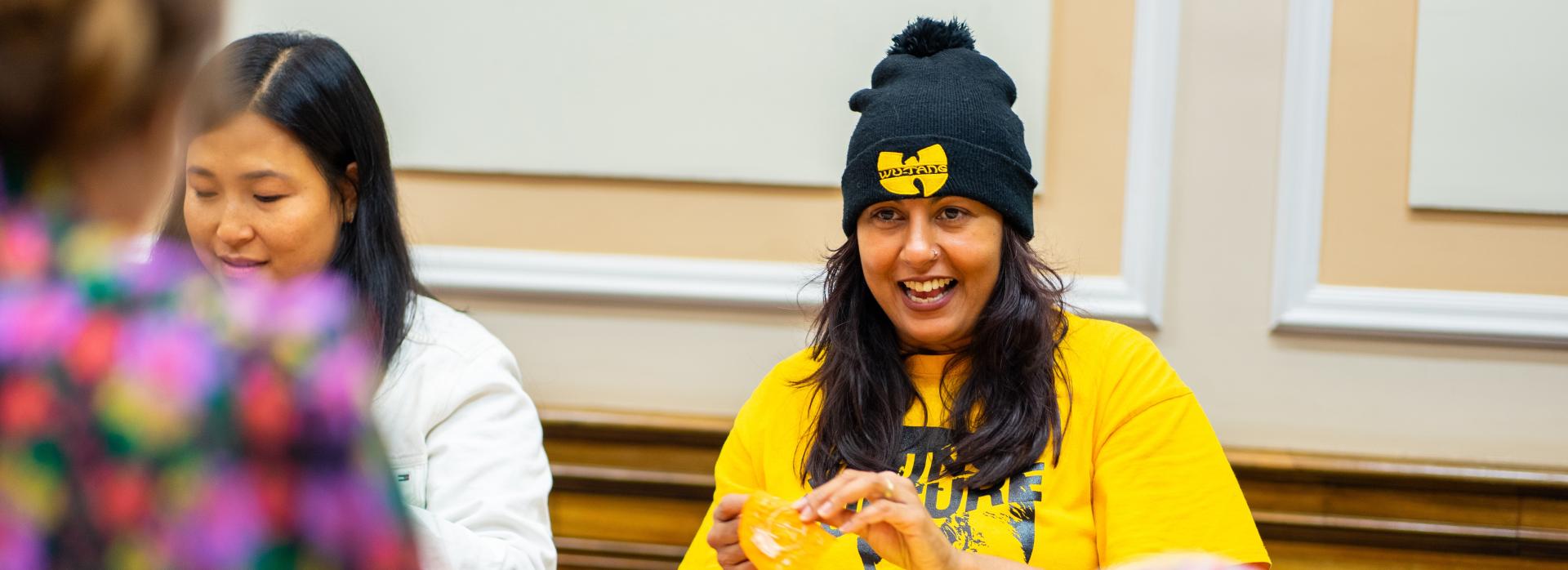 a person in a yellow t-shirt with long black hair and a black beanie sits at a table full of colourful scraps of material