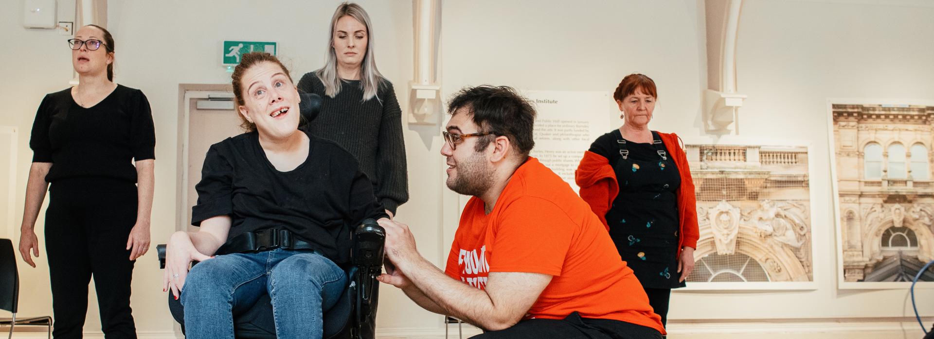 A white woman with brown hair and black t-shirt sitting in a wheelchair and and smiling. A white man with brown hair and glasses, wearing an orange top kneeling beside her, holding her hand. Four people standing behind them.