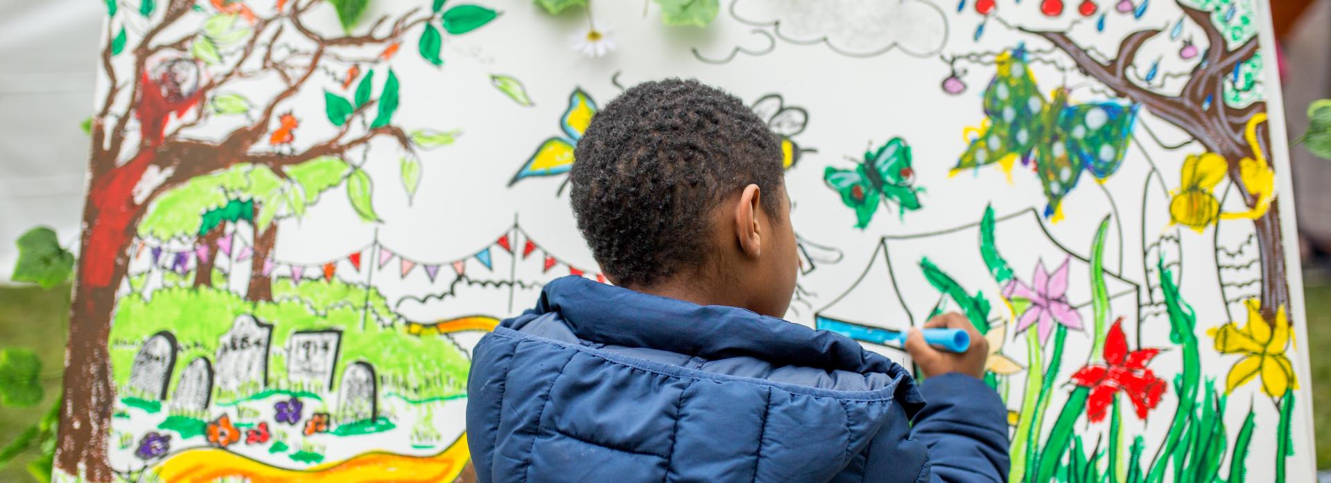 A young black boy wearing a navy blue jacket facing away from the camera painting on a colourful canvas