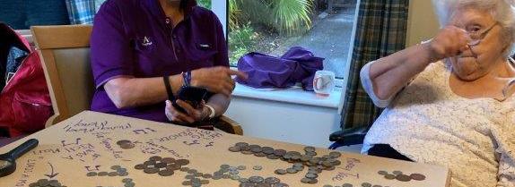 two women, one in a mask, sit at a table with coins all over it