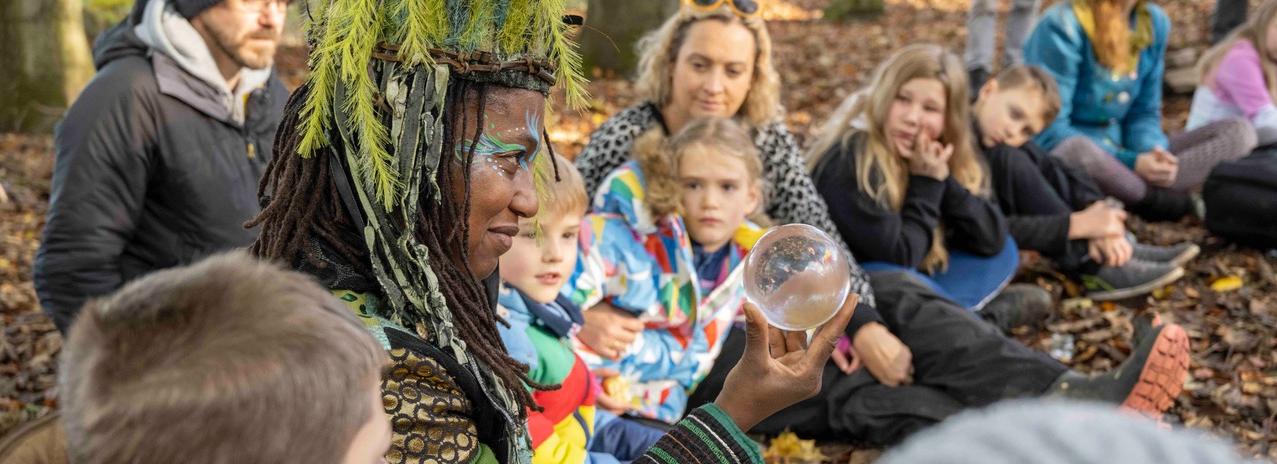 A woman holds up a glass ball, sitting in a circle surrounded by young people and their families in a natural setting