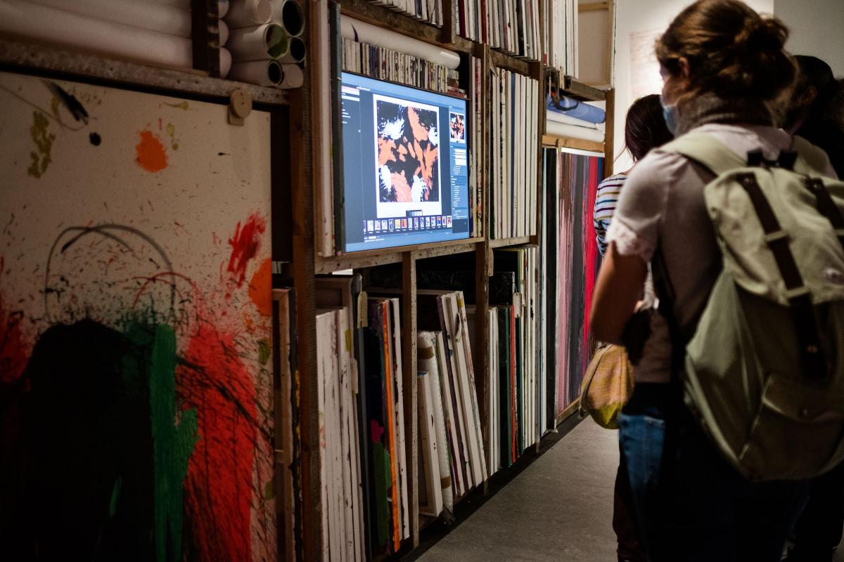 a small group of people looks at a video screen hung in front of many paintings stacked in a storage space