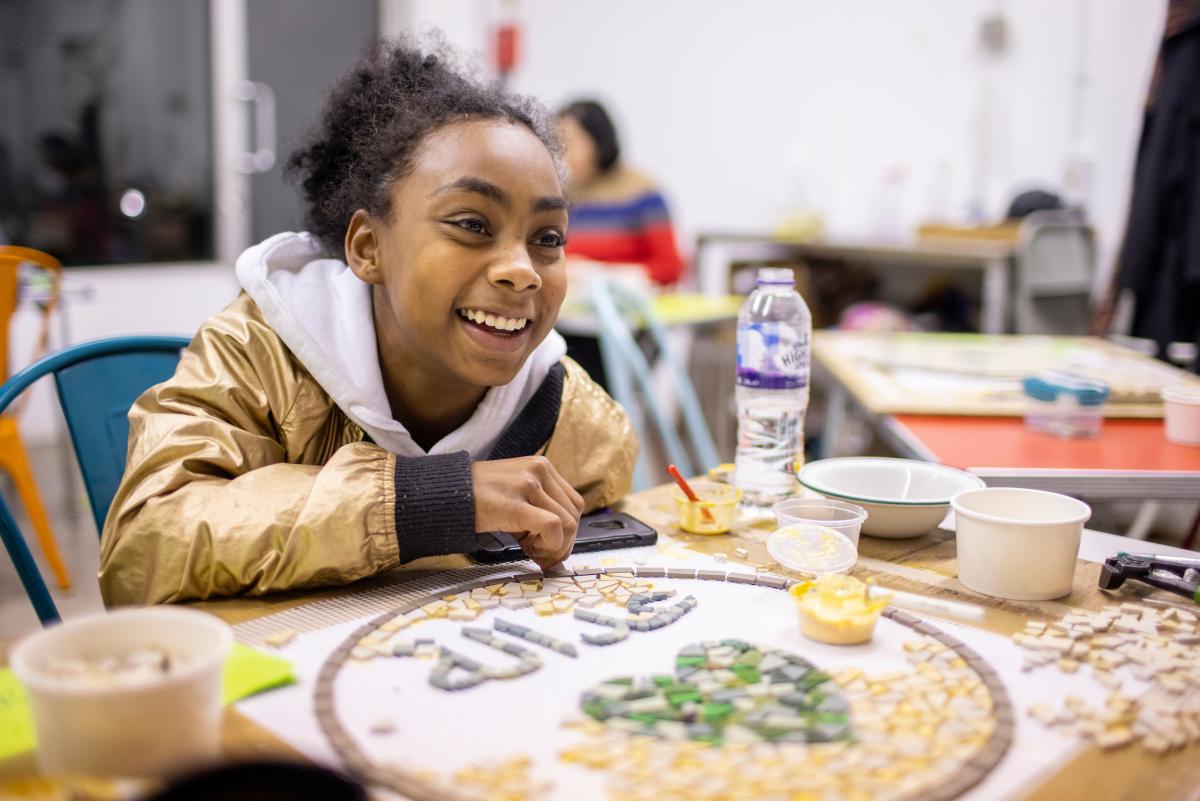 Young girl sits at a table making a mosaic and smiling