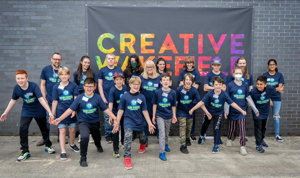 A group of young people and staff stand in front of a wall with the words Creative Wakefield in coloured letters behind them