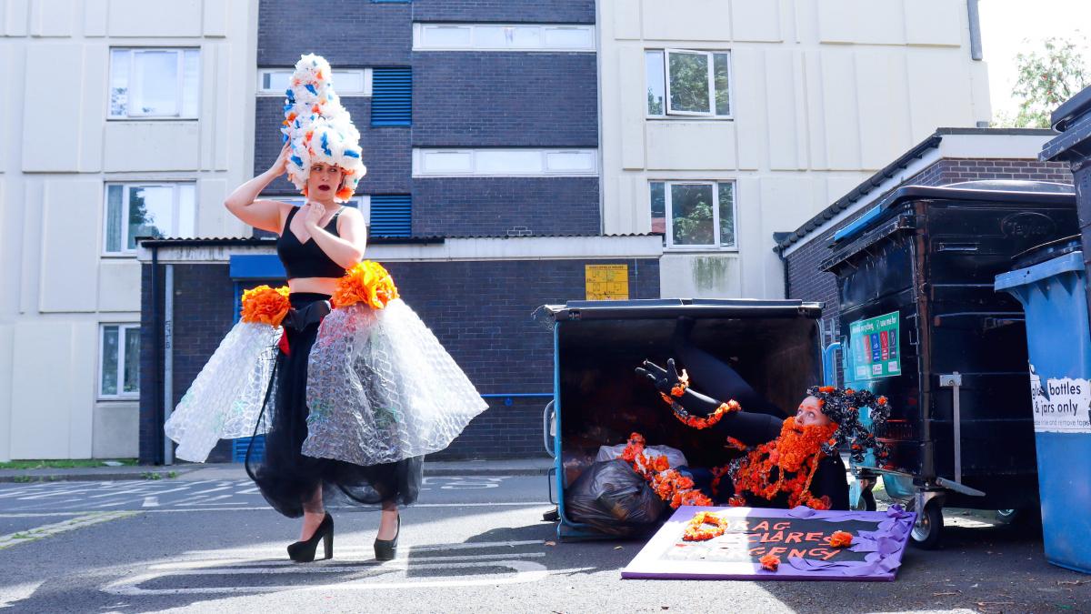 Colour photo, exterior. Kitt, a shaven headed white human, is crawling out of an industrial sized bin with arms outstretched towards Sarah a white human who is looking appalled.  They are both wearing extravagant drag outfits made from garlands of flowers handmade from recycled black, orange, white and blue plastic bags. 