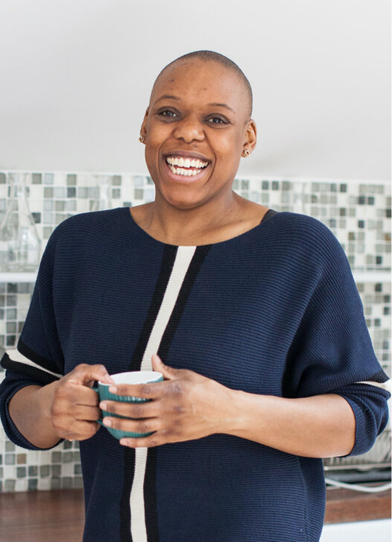 a person stands in a kitchen in front of a tiled wall, holding a cup of tea and smiling