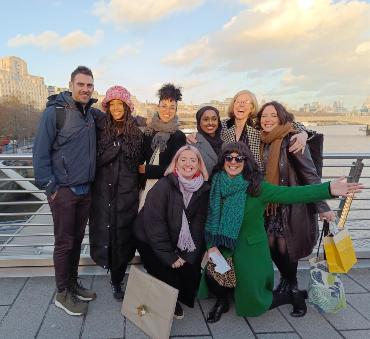 A group of eight people in winter clothing standing on a bridge looking to camera. They are smiling and laughing with arms around each other