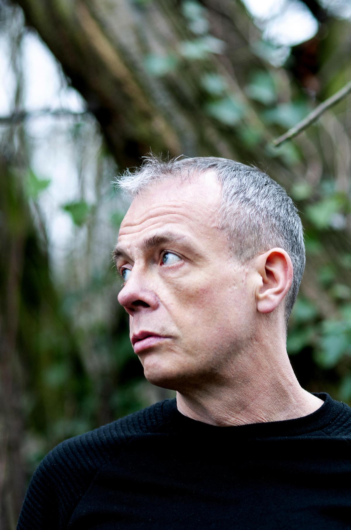 A white male which a closely shaved head looking to his right amongst a tree blurred background