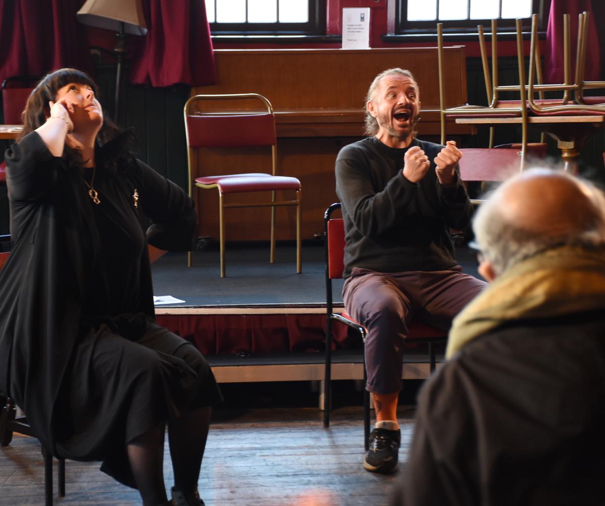 A white male with his hair tied back sitting on a chair in a theatre space with an excitable facial expression alongside a woman wearing all black sitting on a chair looking up towards the ceiling.