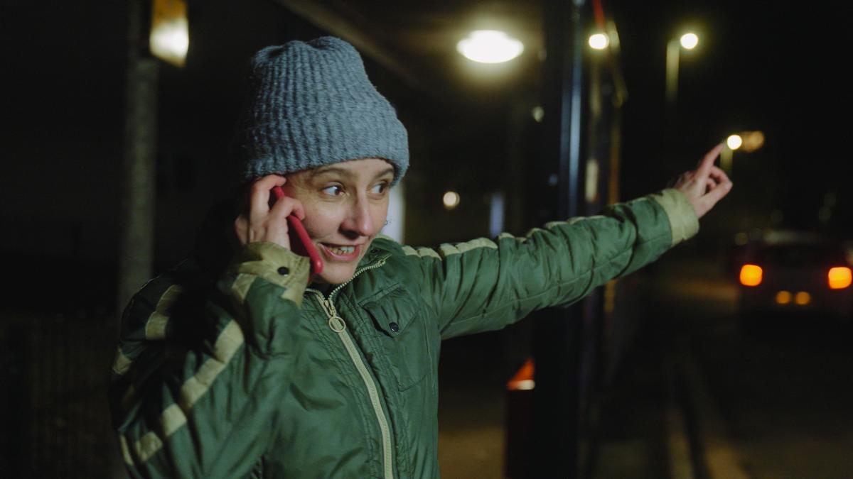 White person wearing a green puffer jacket and a grey wooly hat with their left arm stretched out and holding a red mobile phone to their ear with the other