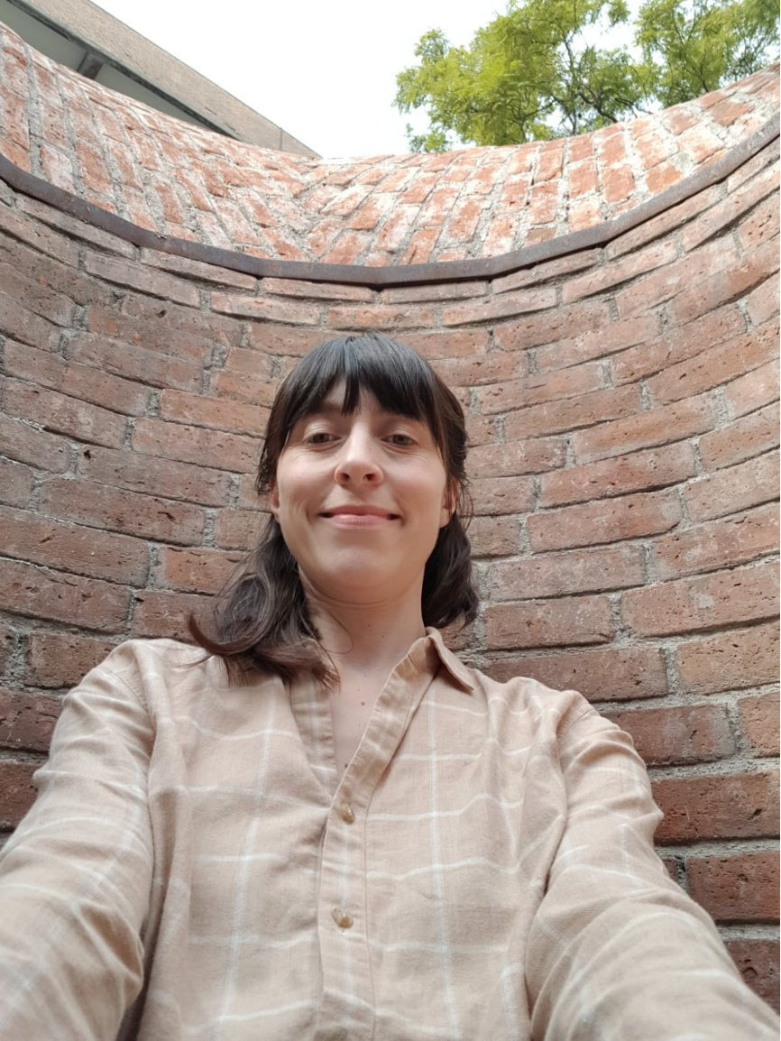 White woman with dark brown hair wearing a chequered beige shirt facing the camera smiling in front of a brick wall.