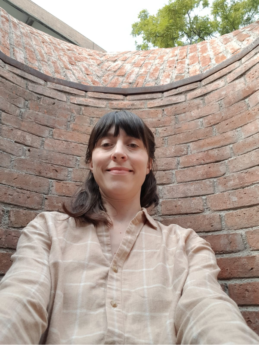 White woman with dark brown hair wearing a chequered beige shirt facing the camera smiling in front of a brick wall.