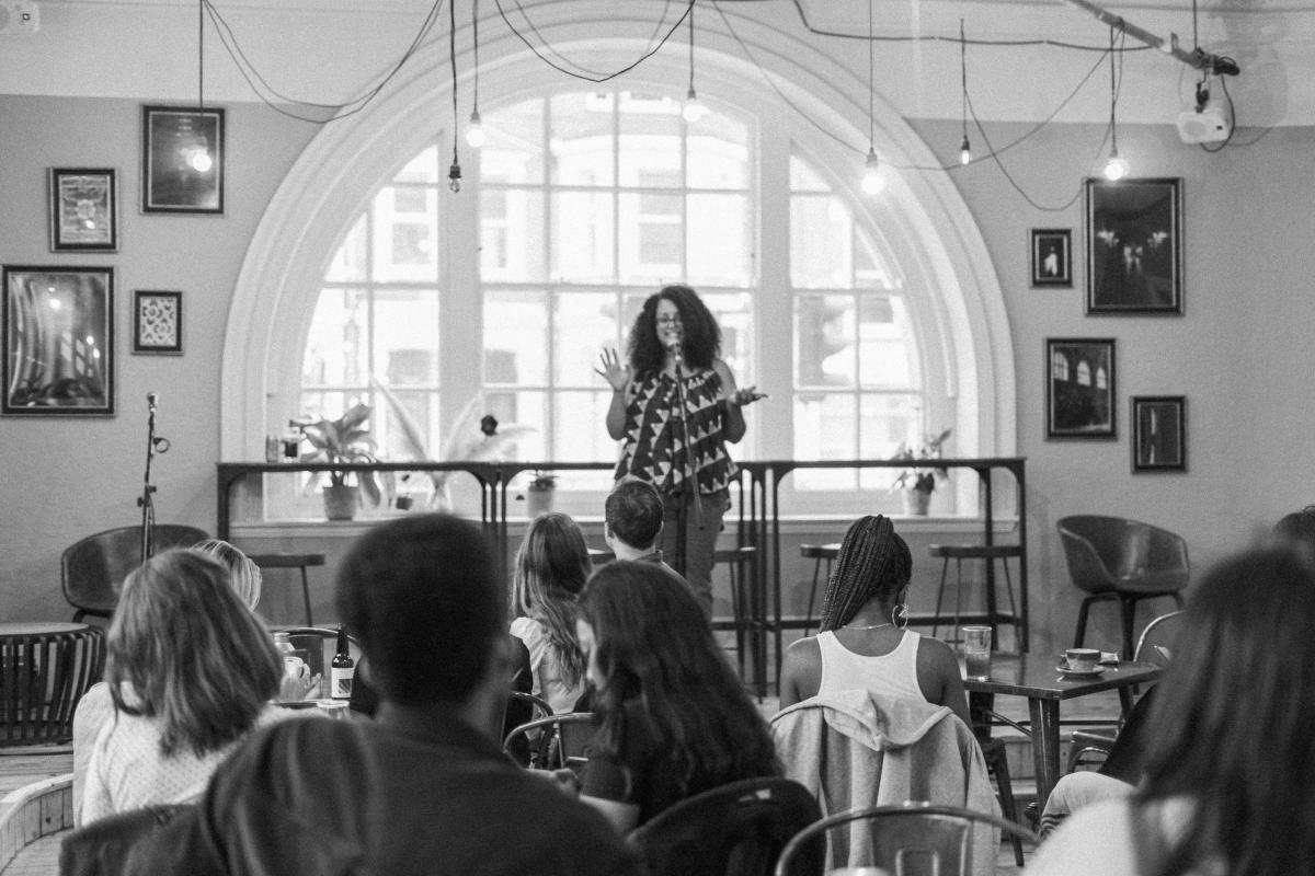 a woman speaks in front of an audience in a brightly lit room. she is standing in front of a large window onto a street.