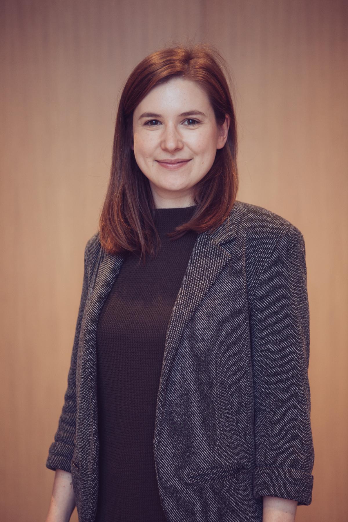 Formal portrait photograph of a woman facing the camera. She has long brown hair and is wearing a grey jacket and black top.