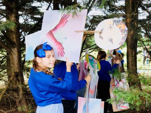 Young girl in a blue school uniform holding on to a piece of artwork suspended in trees, more young people in background doing the same