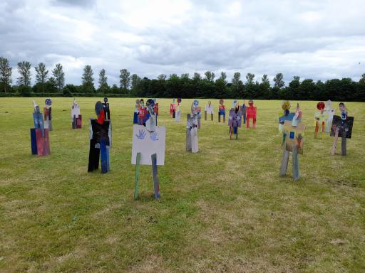 Artworks situated on a field, appear like figures, different colours and creative techniques. Trees in the horizon.