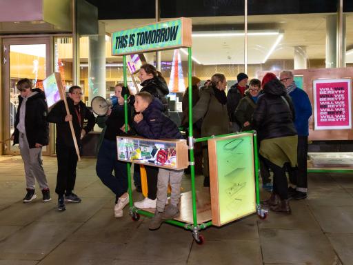 A group of young people standing with placards, loudspeakers and a stand with the words, This is Tomorrow, on the banner. It is nightime with light from the building behind.