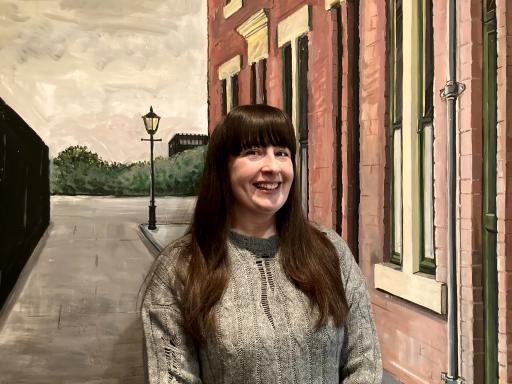 white woman with long brown hair wearing a grey jumper standing in front of a wall with an image of houses and a street