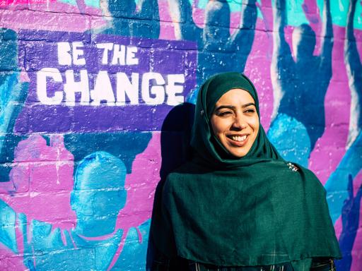 A South Asian woman wearing a dark green coloured headscarf, smiling slightly away from the camera, standing in front of a colourful graffiti wall mural with the words "Be the change" 