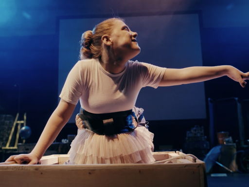 A woman in a pink tutu emerging from a box with her arm held out. She wears a back support