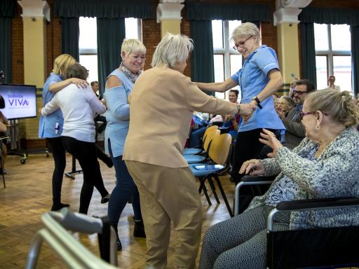 A group of older people and nurses, listening to live music. Some are dancing, clapping and smiling.