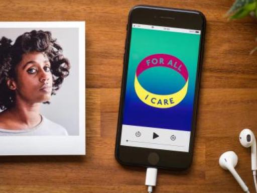 Black iPhone attached to Apple headphones resting on a desk next to an image of a young black woman with a lose curled afro wearing a light grey round neck jumper