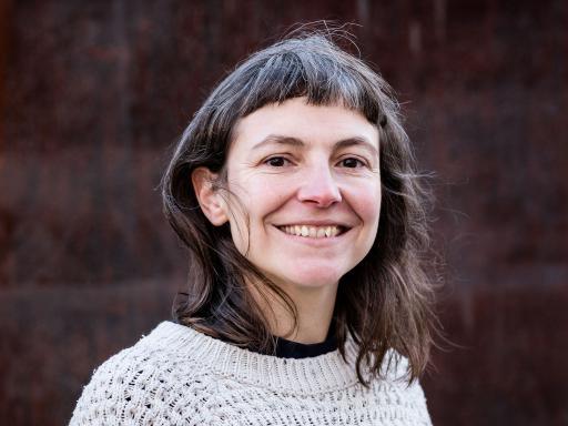 A white woman with shoulder length black hair and a fringe, smiling at the camera in a white knitted jumper in front of a dark brick wall