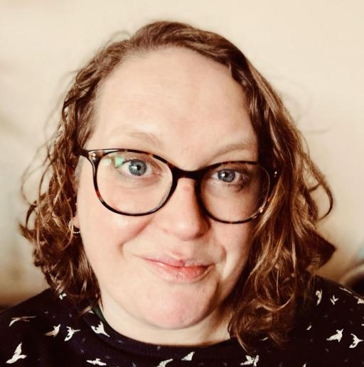 A white woman with shoulder length curly brown hair wearing glasses and wearing a black top with small white birds on, smiling .