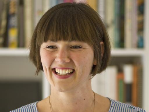 A white woman with a short brown bob wearing a blue dress smiling in front of a bookcase