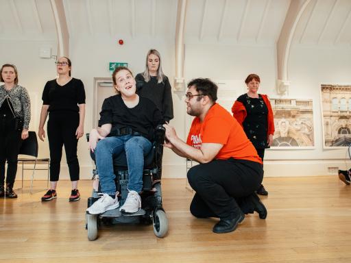 A white woman with brown hair and black t-shirt sitting in a wheelchair and and smiling. A white man with brown hair and glasses, wearing an orange top kneeling beside her, holding her hand. Four people standing behind them.