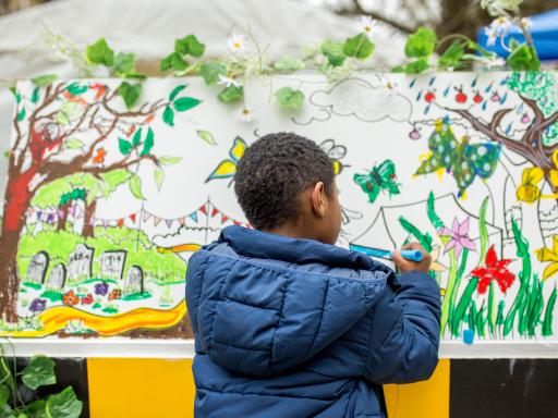 A young black boy wearing a navy blue jacket facing away from the camera painting on a colourful canvas