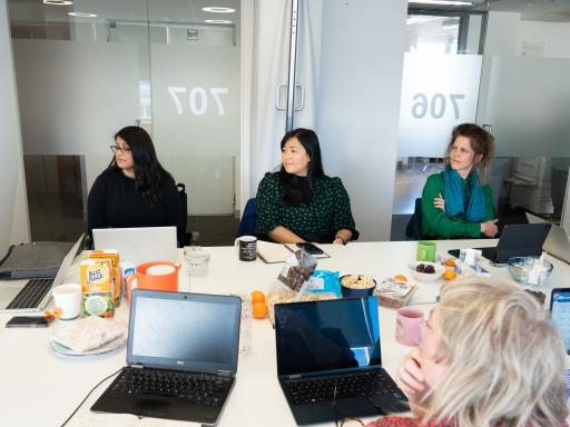 four people sit around a table covered with laptops, snacks and papers