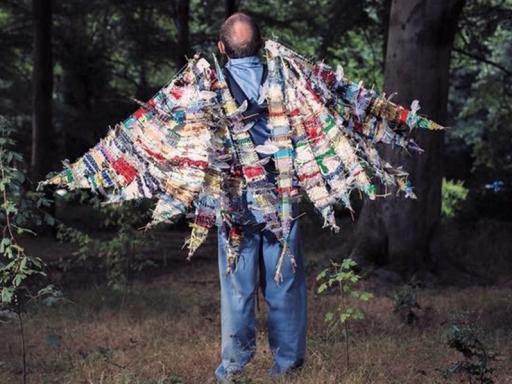 A man facing away from the camera and into a wood. On his back are large wings made of scraps of fabric.