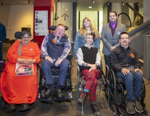 six people, four of whom are using wheelchairs, look directly at the camera smiling in a room in a museum