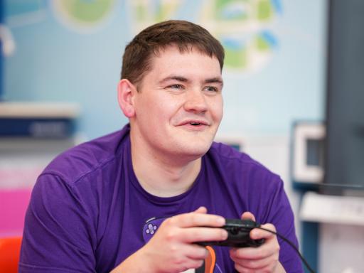 Young man with black short hair, wearing a purple t-shirt holding a console control pad and smirking