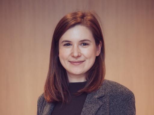 Formal portrait photograph of a woman facing the camera. She has long brown hair and is wearing a grey jacket and black top.