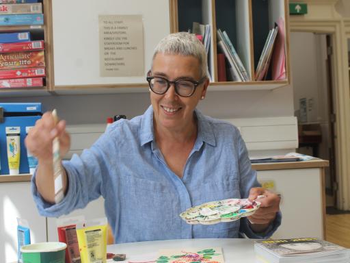 A white woman with short blonde hair and glasses, holding a paintbrush and palette