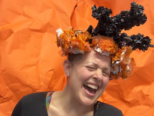 A white shaven headed human has a big smile wears a headdress made of huge multi coloured flowers and a black long sleeved shirt on which white and orange text reads “ queer crip craft power”