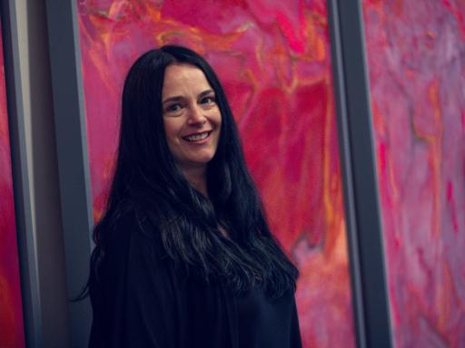 A dark haired white woman against a pink background, smiling to camera