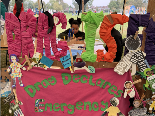   	 A portrait image. A pink fabric banner that reads 'Drag Declares Emergency Glean Hope' in green fabric letters. Paper mythical beasts surround the banner, created by workshop attendees. The banner is attached to a wooden sign that reads 'Drag is Folk' 