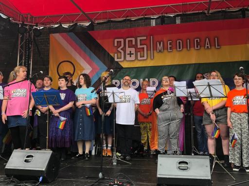 photo of a choir standing on a stage with a large pride banner behind them