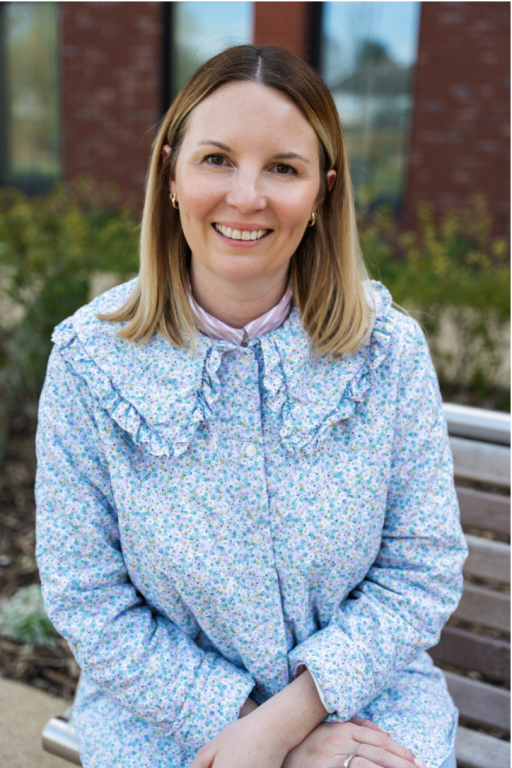 A woman with brown shoulder length hair wearing a blue frilly dress with her hands in her lap smiling at the camera